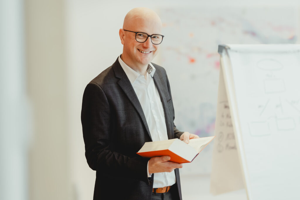 Christoph Becker lächelt hat eine Brille auf und ein Buch in der Hand